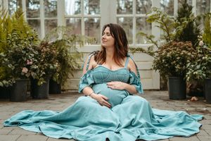 Expectant mother in flowing turquoise dress sits peacefully in a sunlit greenhouse surrounded by lush greenery.