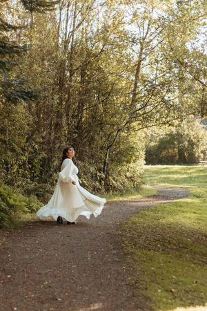 A person in a flowing white dress sits gracefully on a winding dirt path surrounded by autumn trees and golden light.