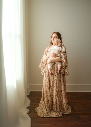 A newborn baby swaddled in white is cradled in a woman's arms wearing a flowing floral dress against a neutral backdrop.