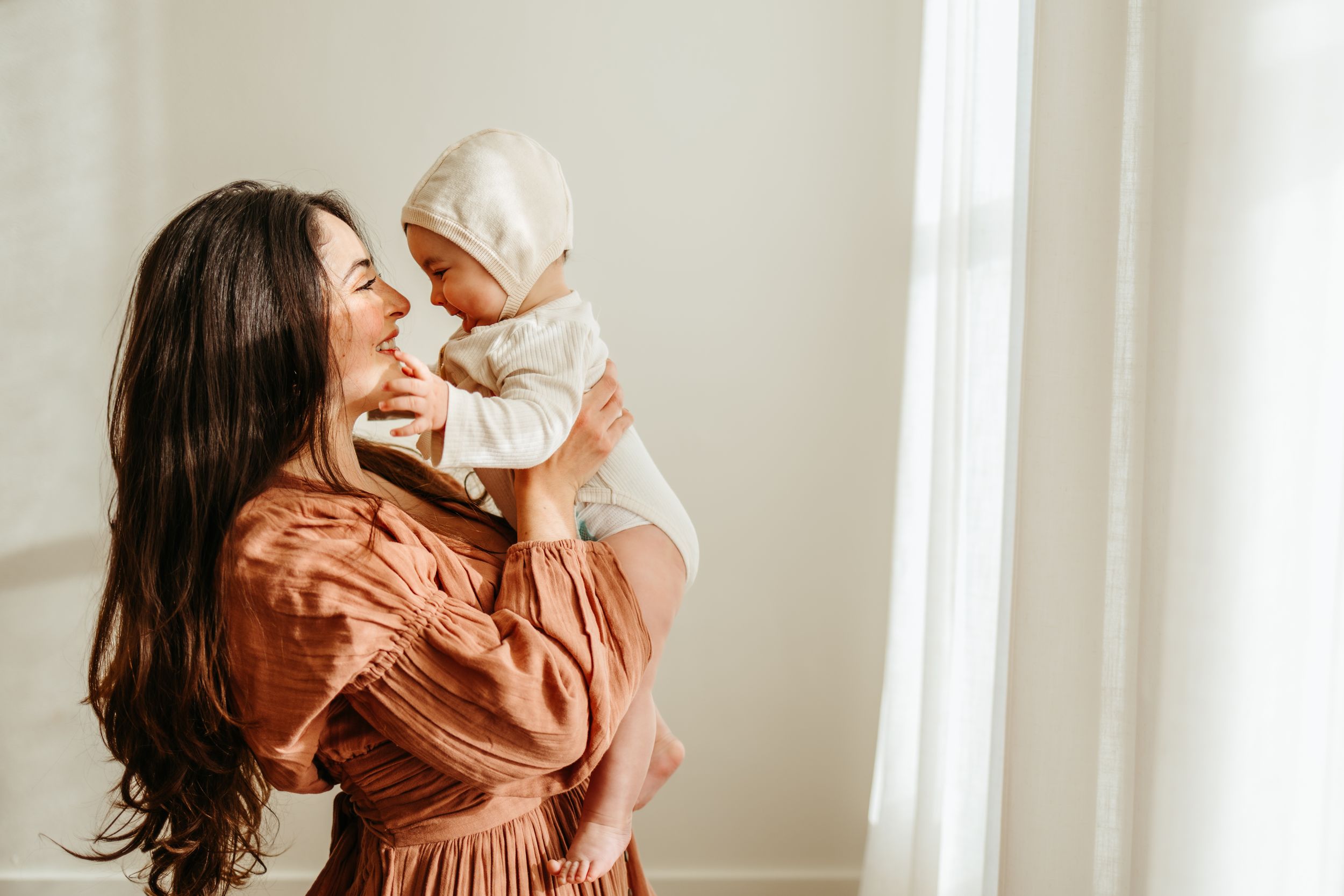 A mother in a rust-colored dress shares joyful moments with her baby in a bright, airy room.