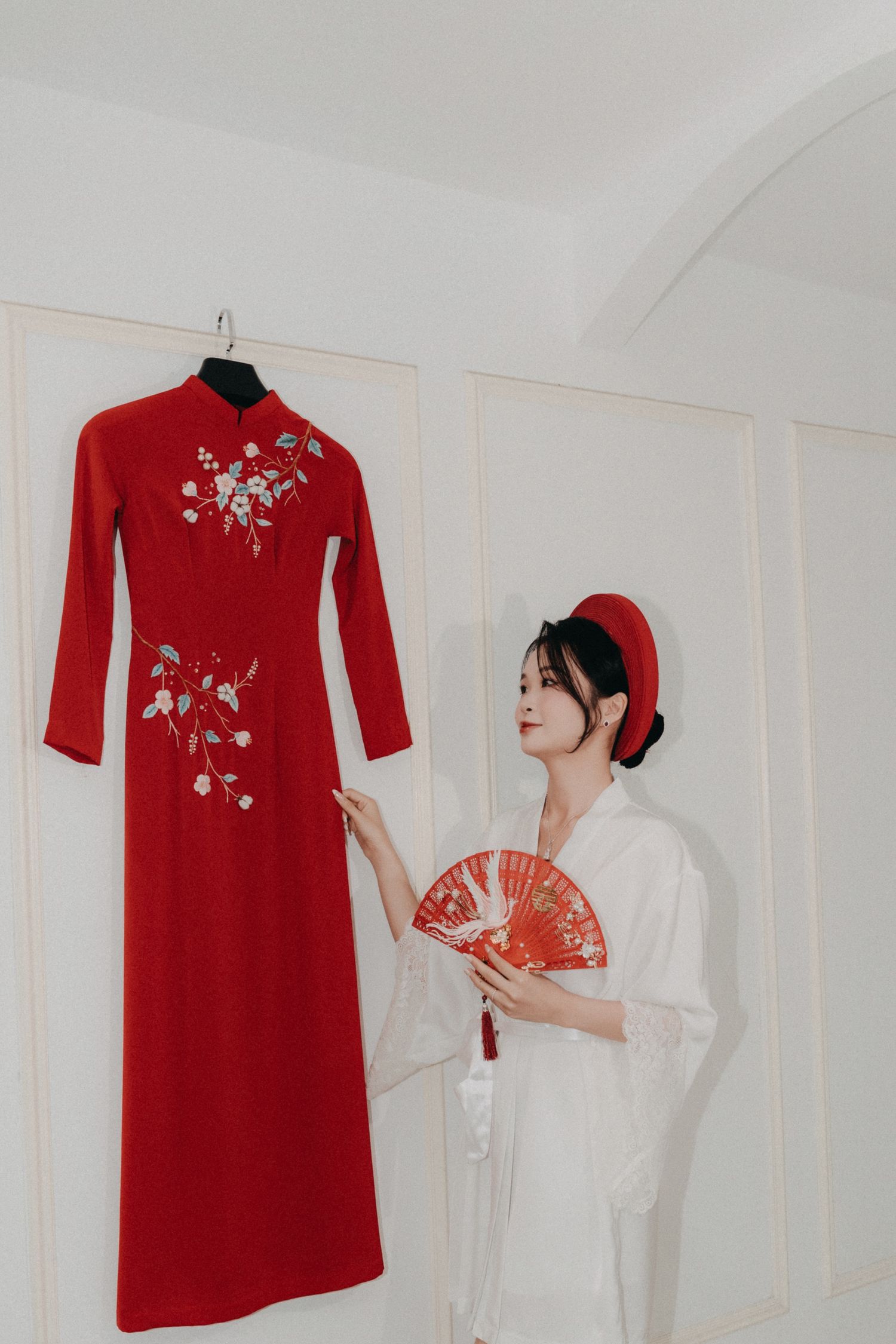 Bride in white robe admires embroidered red ao dai gown hanging on wall before ceremony.