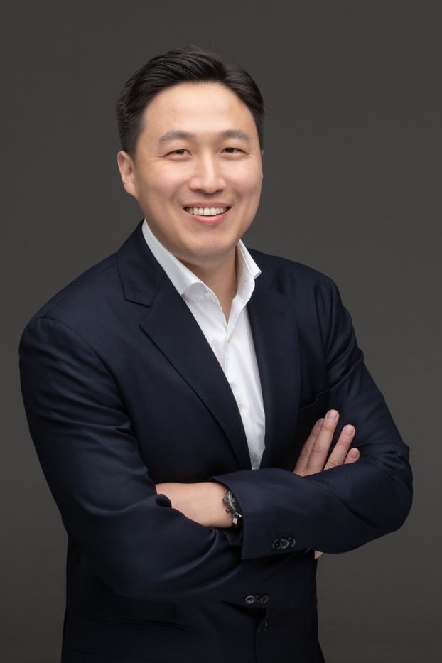 Professional business headshot of a person in a dark suit and white shirt smiling against a gray background.