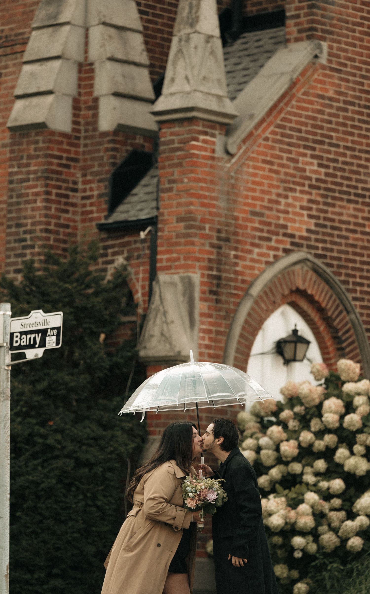A couple shares an intimate moment under an umbrella near a Gothic brick church with hydrangeas.