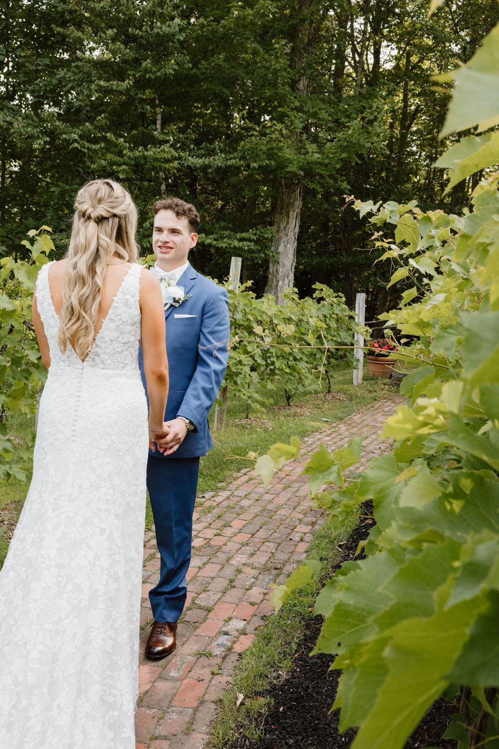 A couple in wedding attire walks along a brick path surrounded by lush greenery in a garden setting.