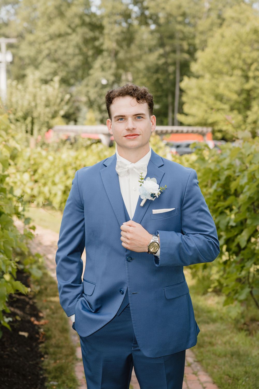A groom in a fitted blue suit stands in a garden setting for formal wedding portraits.