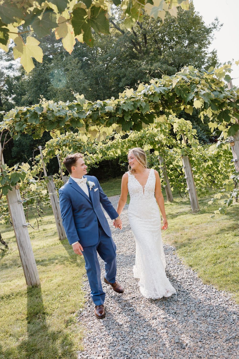 A romantic couple walking through a sun-dappled vineyard with grapevines and gravel path on their wedding day.