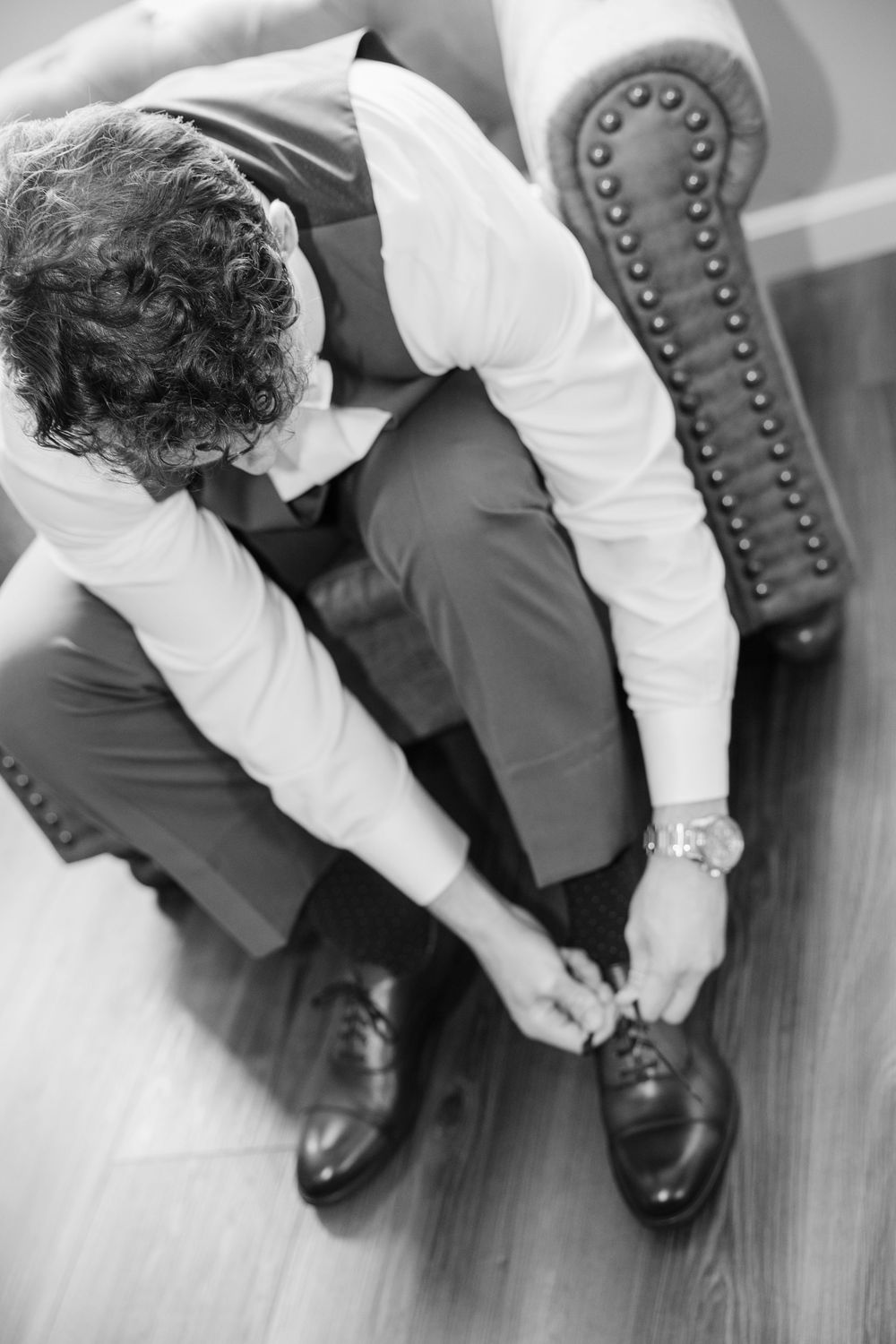 Black and white image of a person tying dress shoes while seated on a studded leather chair.