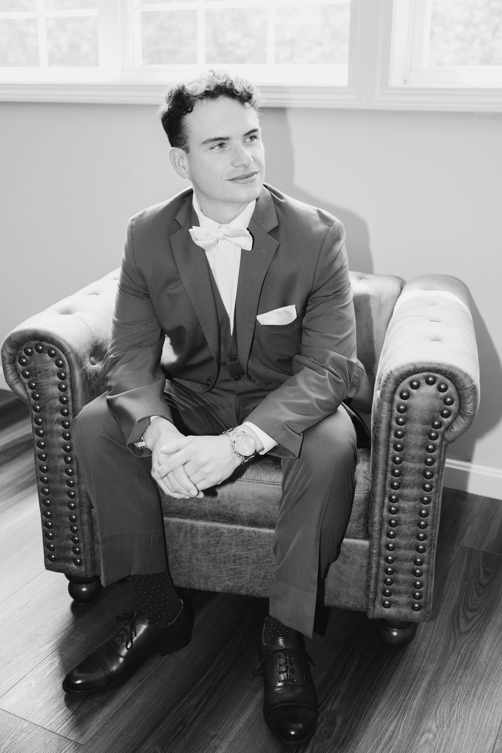 Person in dark suit sits on an armchair in contemplative pose with hardwood floor reflecting ambient light.
