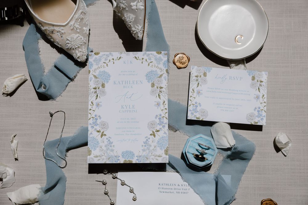 Elegant wedding invitation suite with delicate blue floral designs and silk ribbons arranged on gray surface.