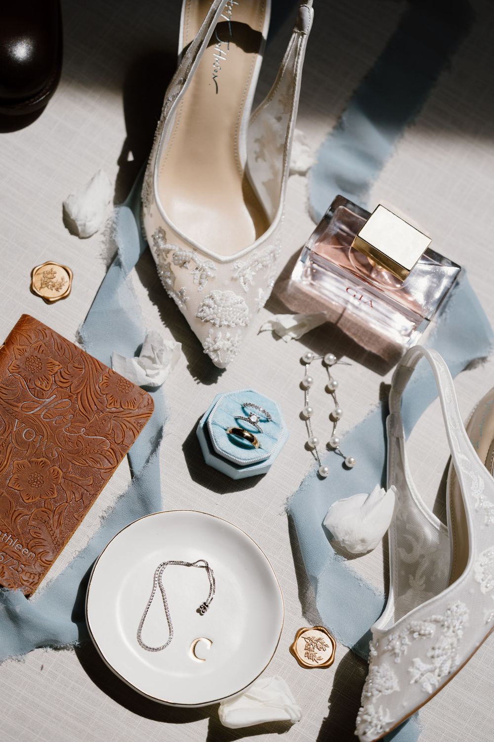 Wedding accessories including elegant white heels with floral details, jewelry pieces, and vintage brown leather accents laid out elegantly.