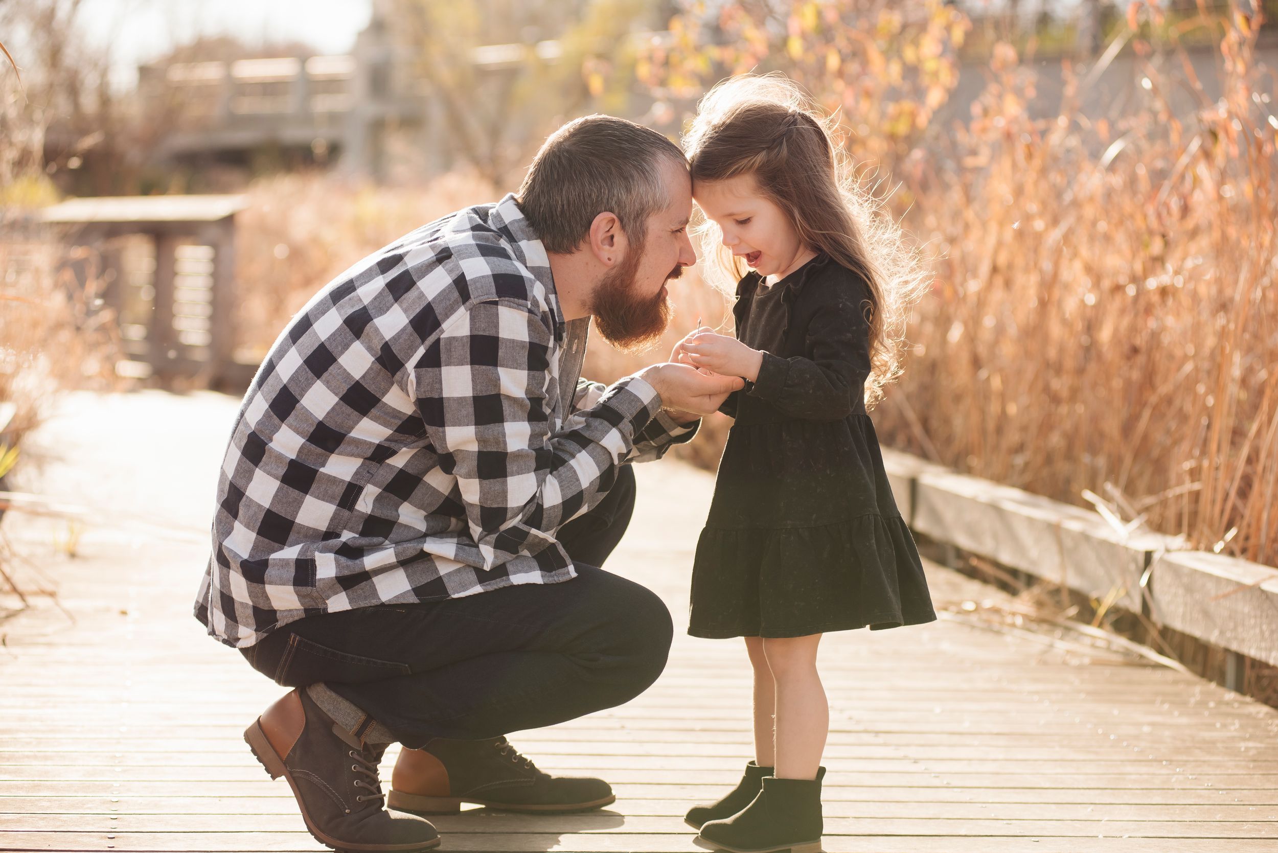 Father kneeling on wooden boardwalk holding hands with young daughter in black dress during outdoor family photo session.
