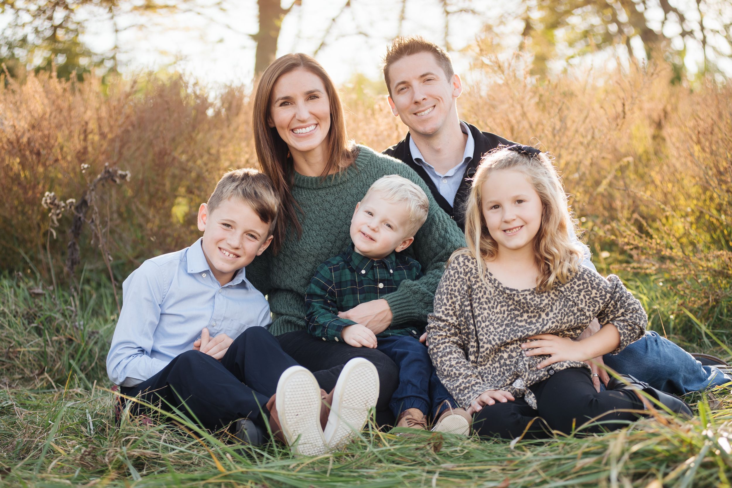 Happy family of five sitting in green grass with autumn foliage, parents with three young children smiling outdoors.
