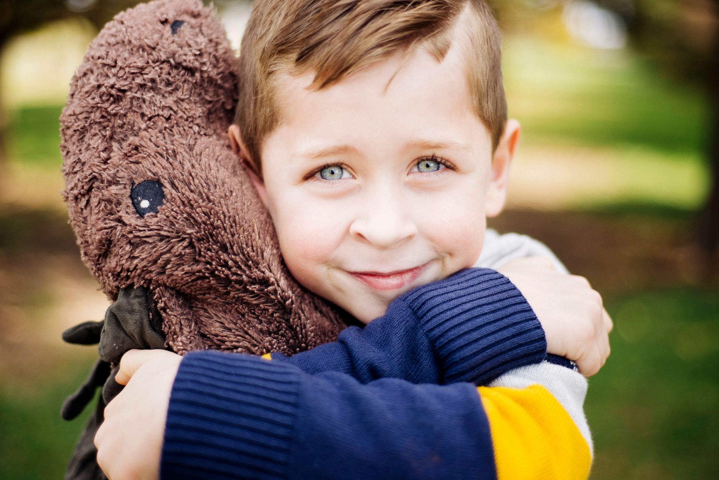 Adorable blue-eyed boy hugging a large brown stuffed animal toy while smiling outdoors in a park.
