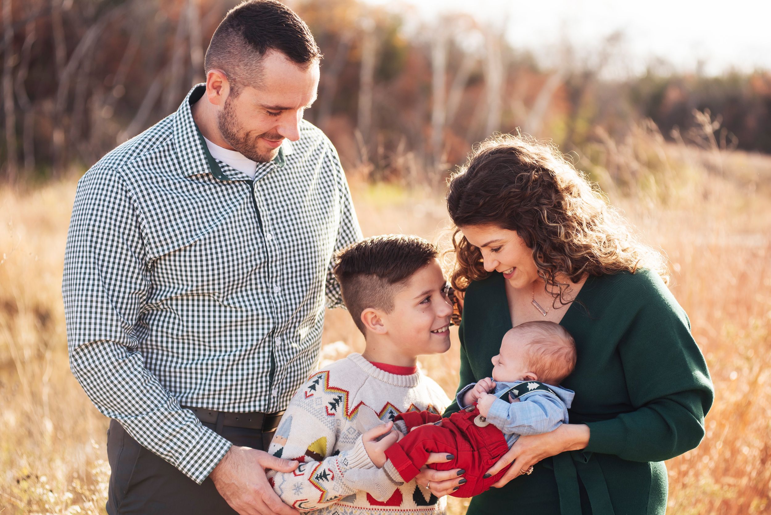 Family of four outdoors in golden field, parents smiling at newborn baby while young son stands between them.