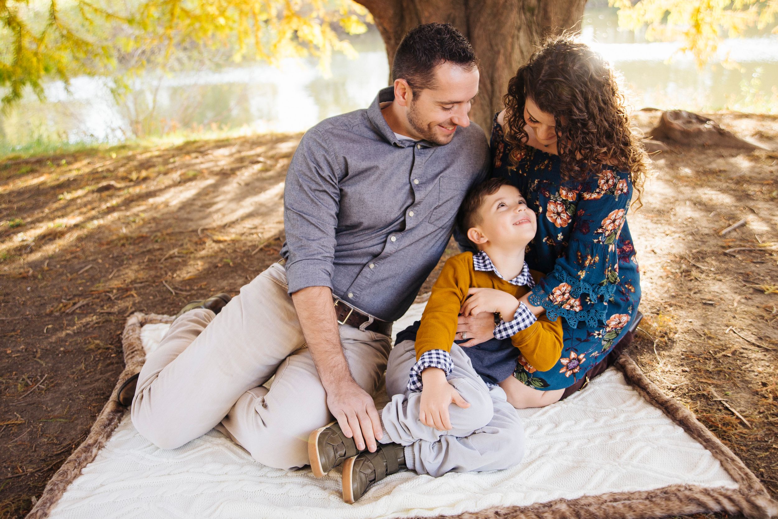 Happy family of three sitting on blanket under tree, parents cuddling laughing toddler son in autumn park.
