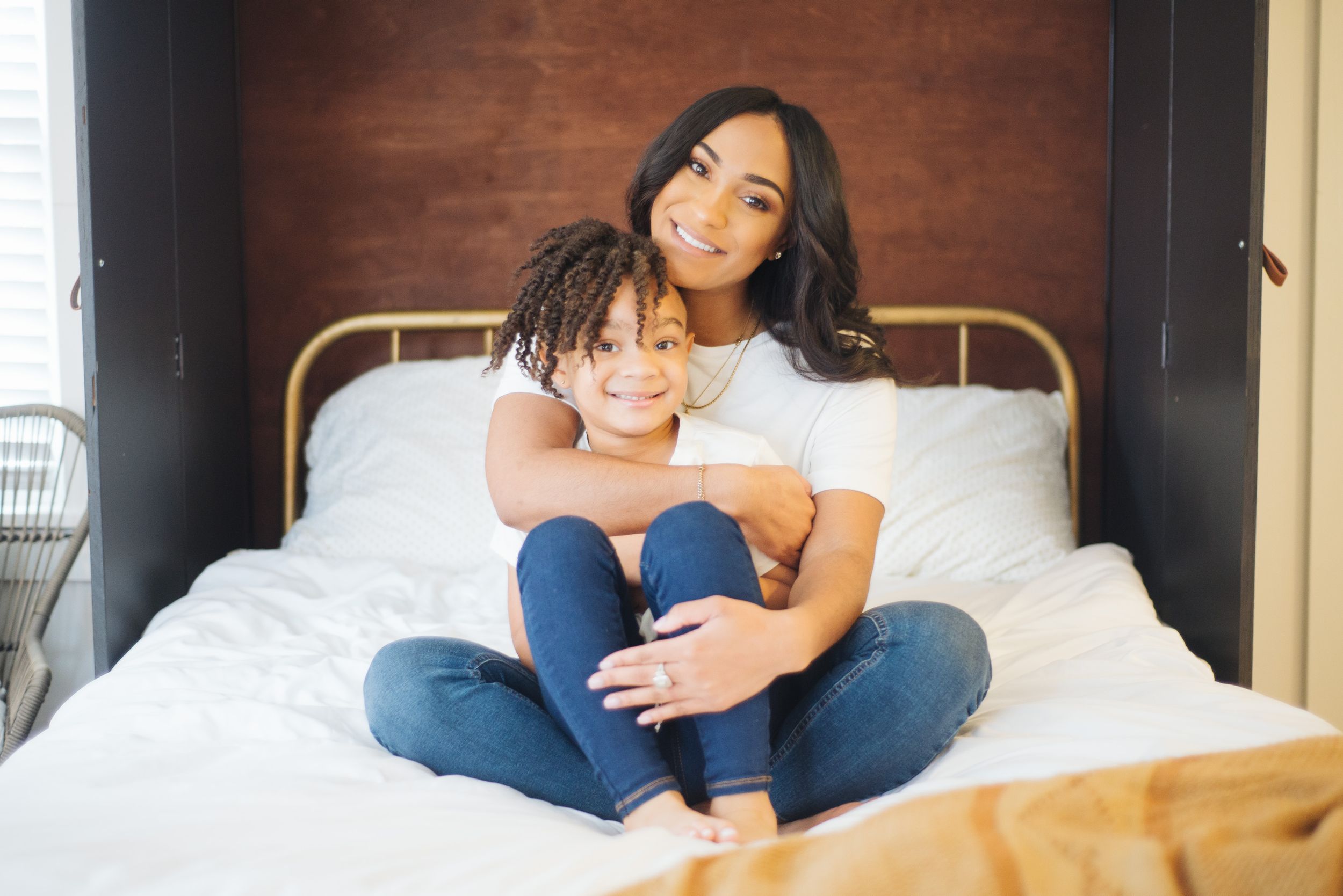 Smiling woman and child sitting together on a white bed in a cozy bedroom.