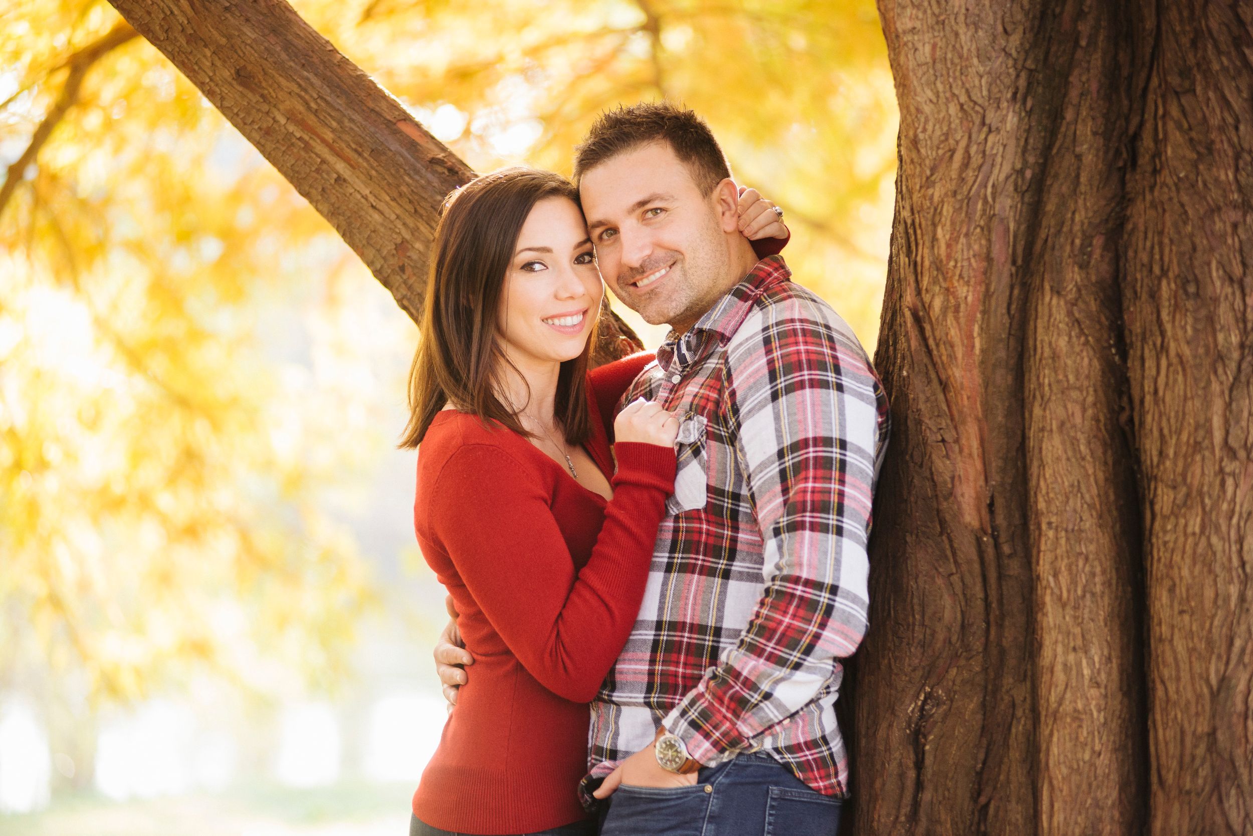 Couple embracing and smiling by a large tree with golden autumn foliage in a park.