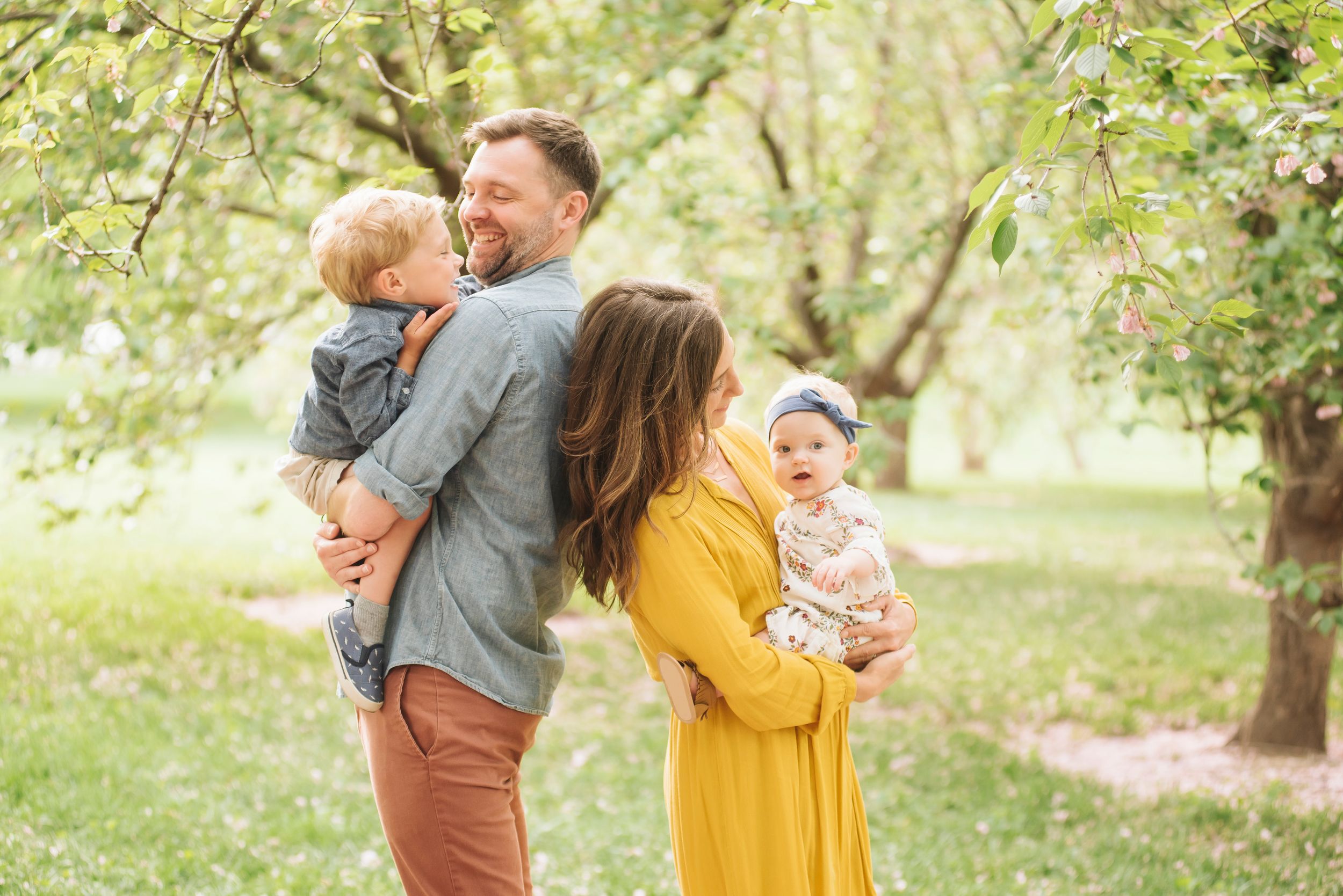 Young family of four sharing a tender moment under blooming trees, mother in yellow dress holding baby girl.