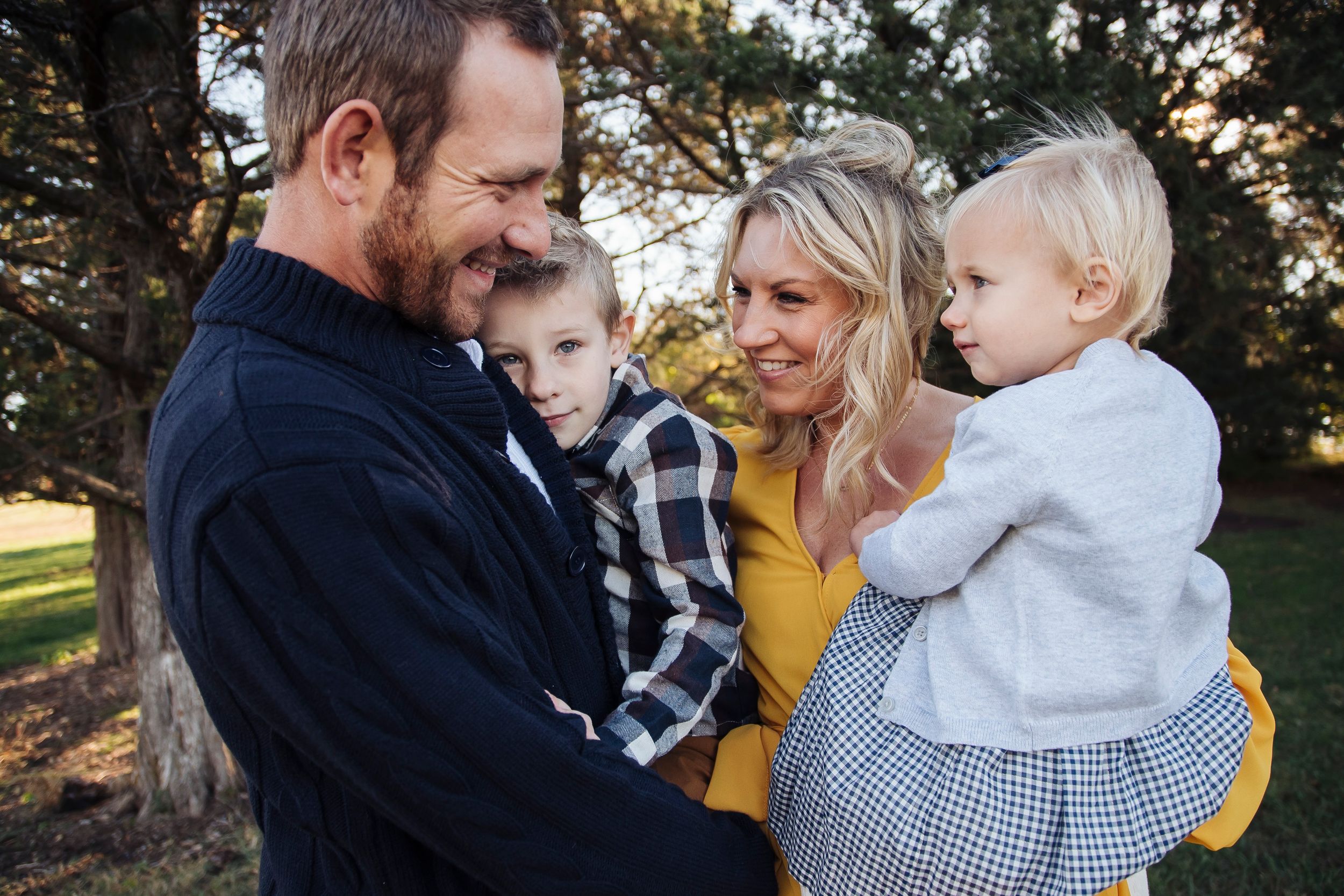 Smiling family of four outdoors in park, parents holding two young children while laughing together.