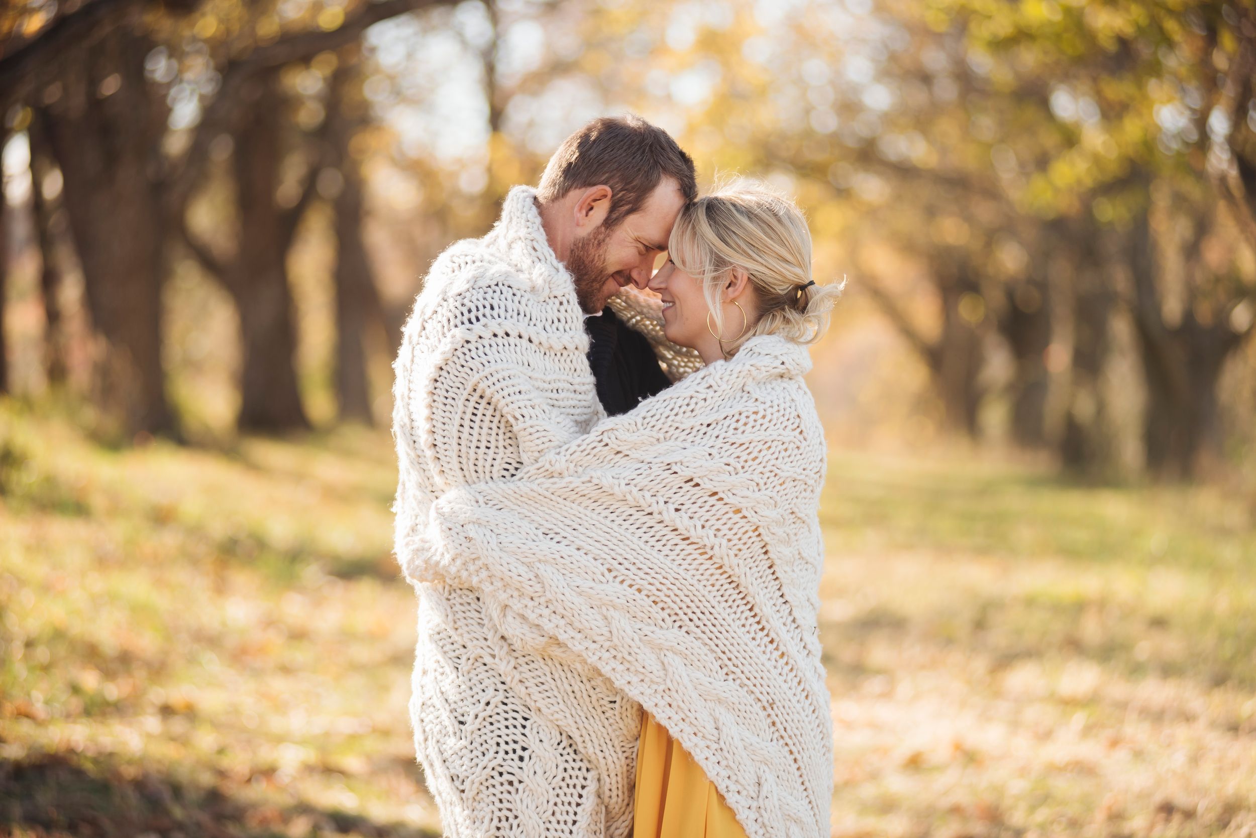 Romantic couple wrapped together in chunky knit blanket touching foreheads in golden autumn forest setting.
