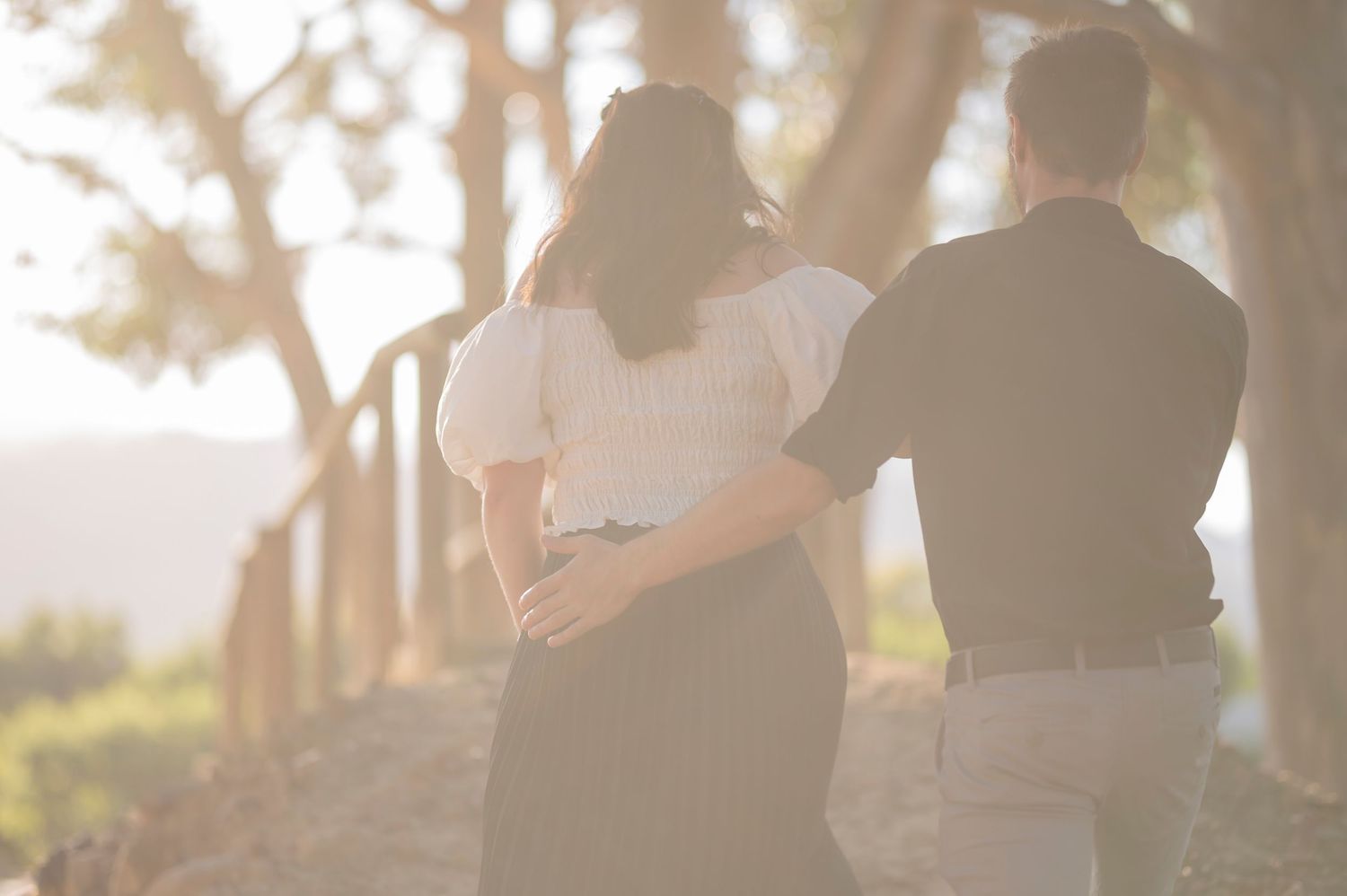 A couple walking together along a sunlit path through trees during golden hour on a hazy afternoon.