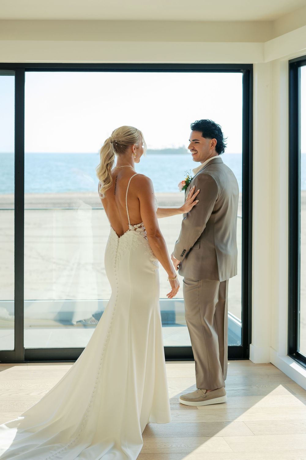 A romantic wedding couple stands by large windows overlooking the ocean in a modern white room.