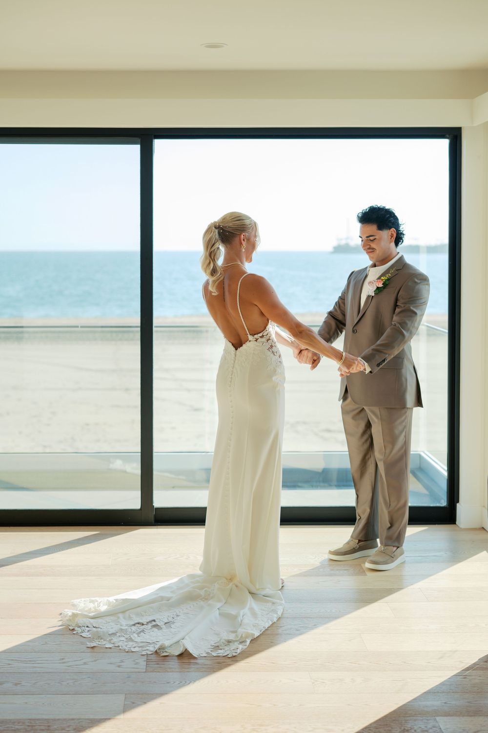 A romantic first look moment between a couple in front of large windows overlooking the ocean.
