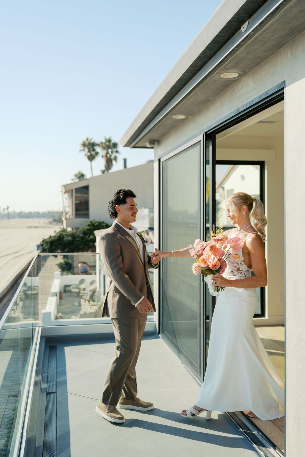 A couple in wedding attire dancing on a sunny balcony with glass railings and modern architecture.
