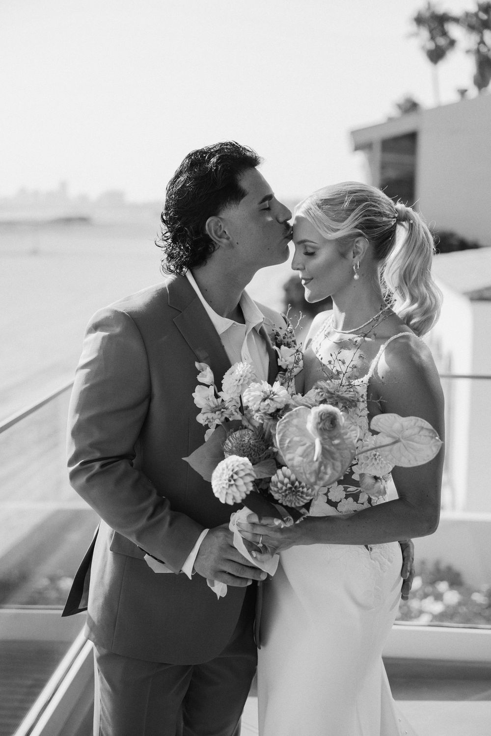 Newlyweds share intimate moments while holding a bouquet of pink and coral flowers on a bright sunny terrace.