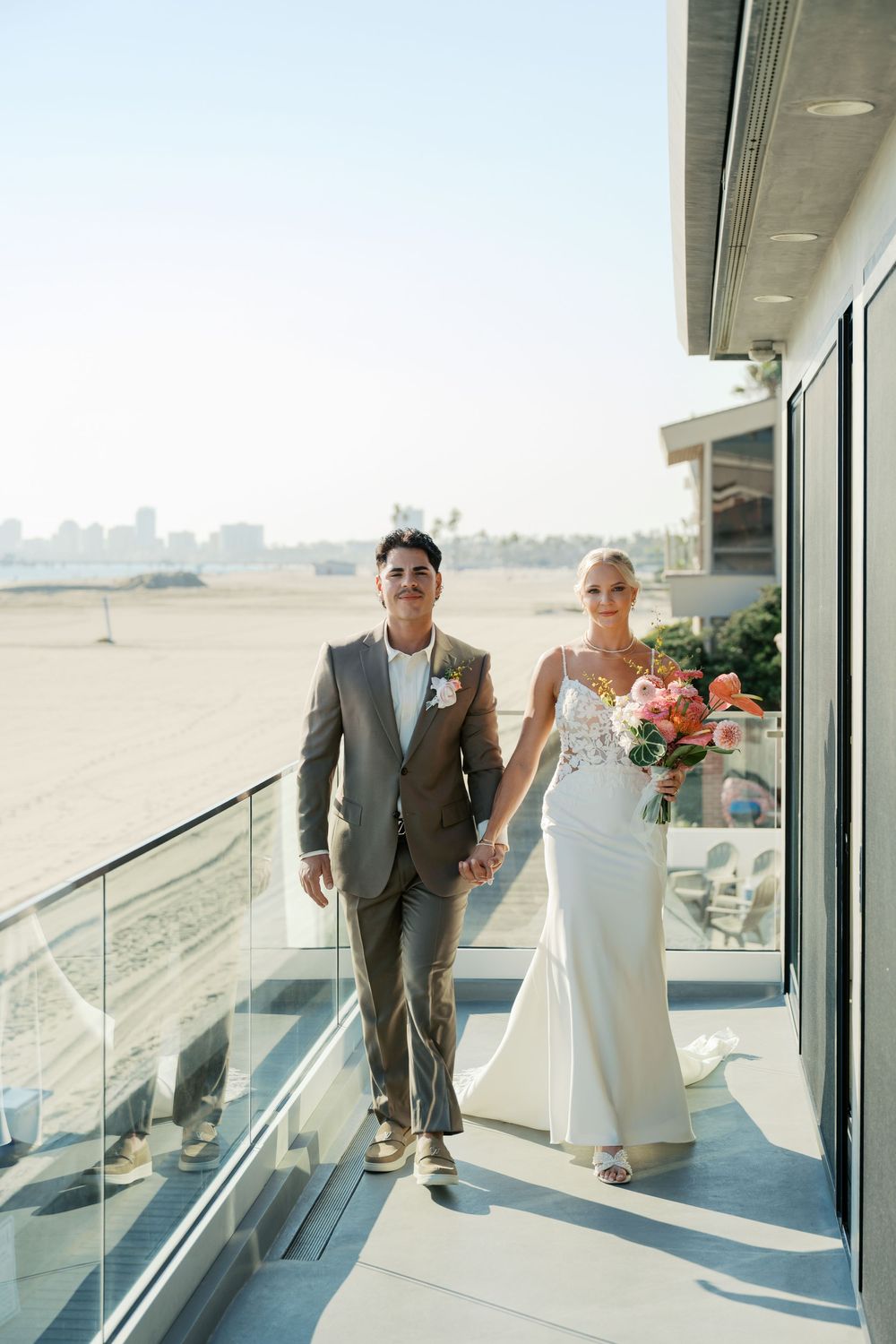 A couple in wedding attire walks along a modern glass-railed balcony with ocean views.