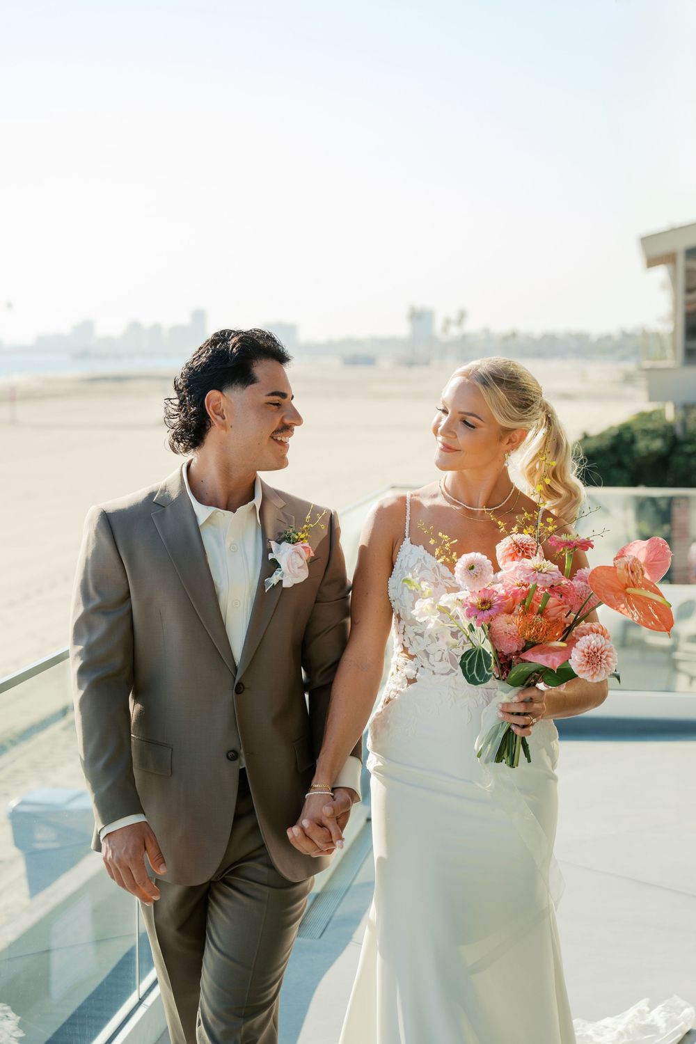 Bride in white dress holds coral flower bouquet as couple shares romantic moments on a sunlit balcony.
