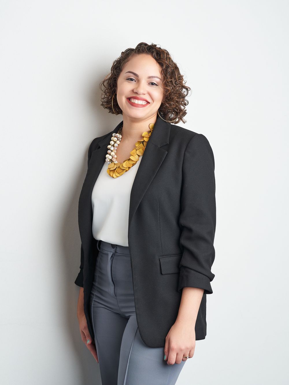 Professional studio portrait showing a person in a black blazer, white top and statement necklace against light background.