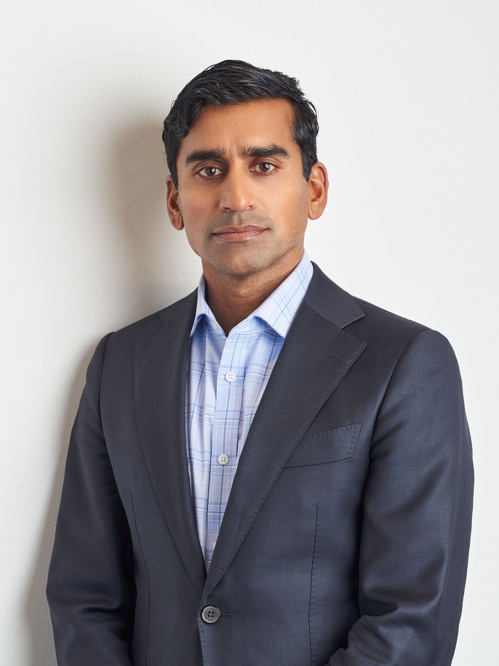 Professional man in dark suit and light blue dress shirt poses for headshots against white background.