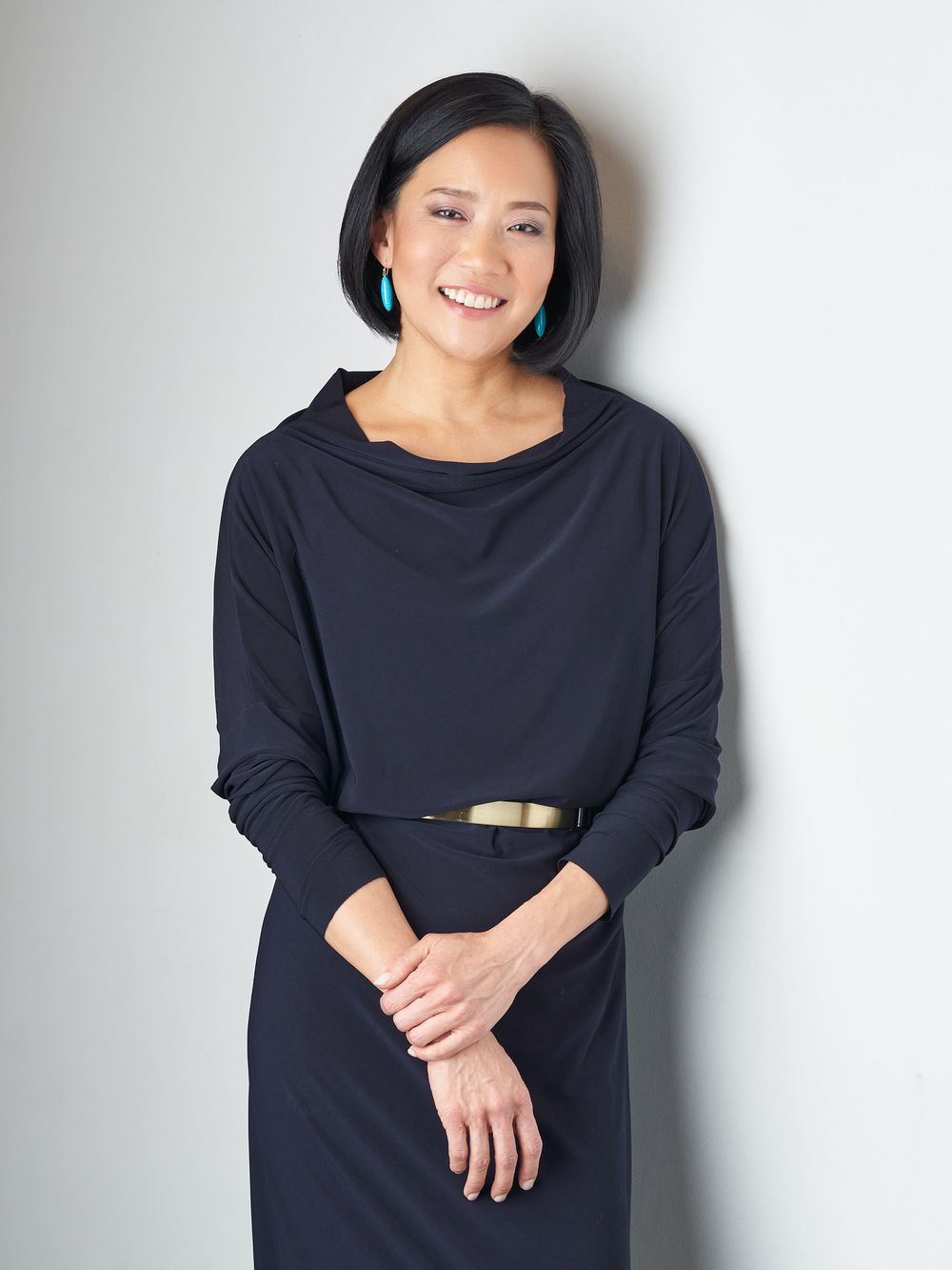 Woman in a navy cowl neck dress with turquoise earrings smiles against grey background.