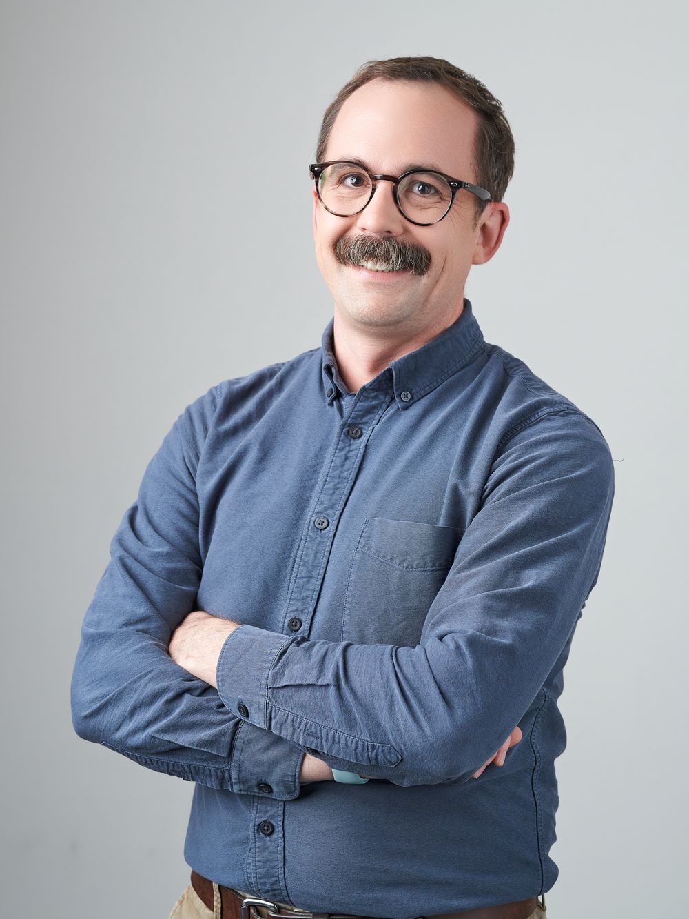 Smiling person in glasses and blue denim shirt poses with arms crossed in studio against light gray background.