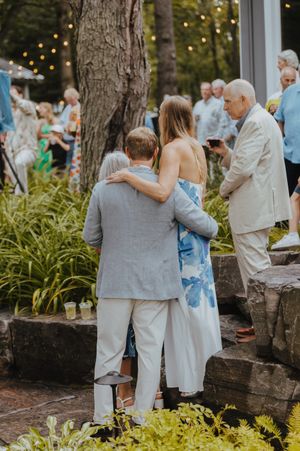 Wedding guests in light colored attire embracing near landscaped garden area.