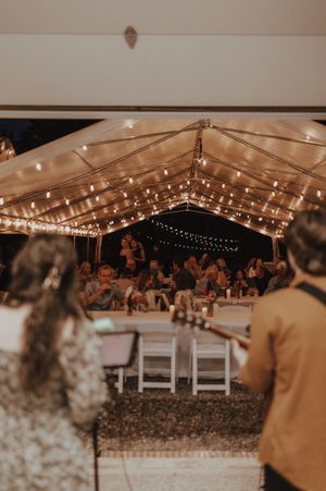 A wedding reception tent illuminated with warm string lights and filled with guests at round tables.