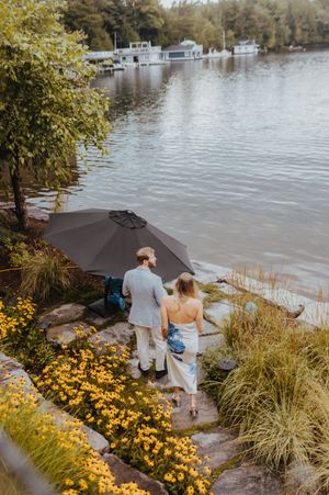 Couple walking along a lakeside path with yellow flowers and boats in the background.