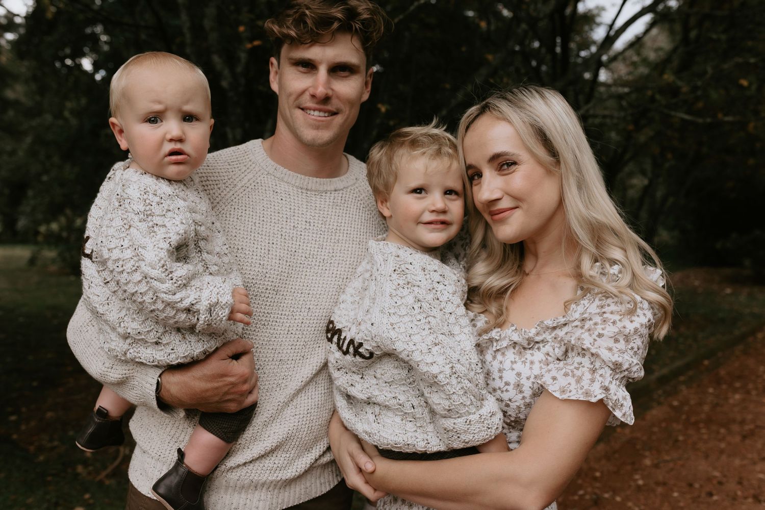 A family wearing coordinated beige and white outfits poses together outdoors in a natural setting.