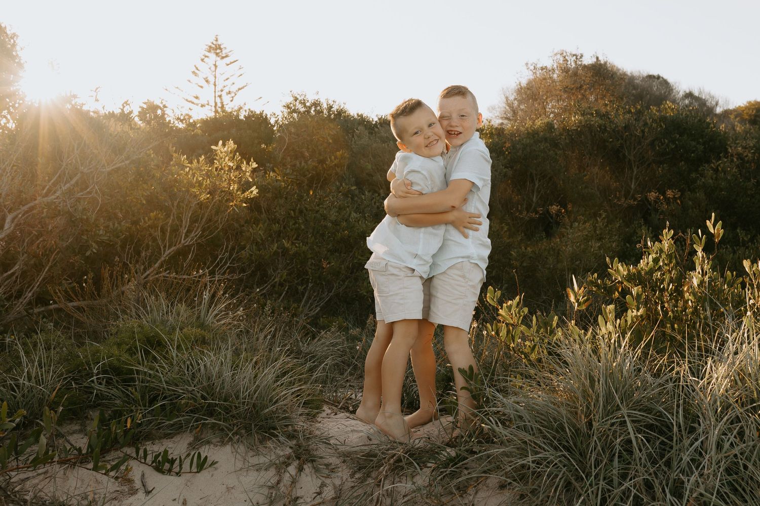 A couple embraces during golden hour sunset on a grassy hillside with warm backlighting filtering through trees.