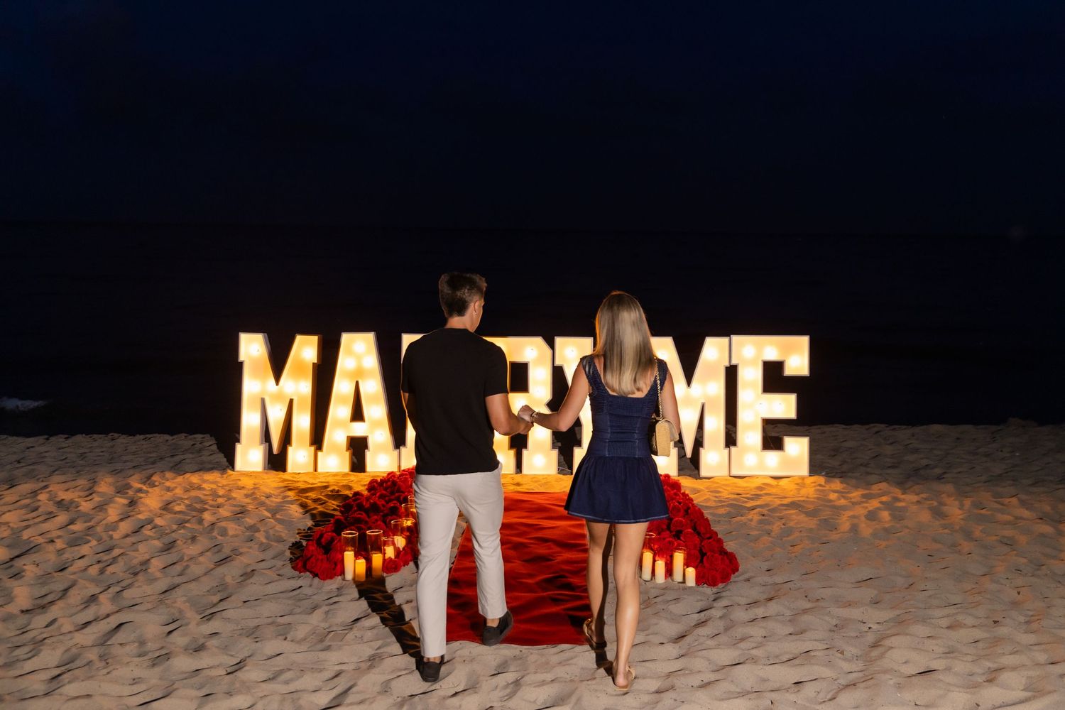 A romantic beach proposal setup at night with illuminated letters and red rose petals forming a path.