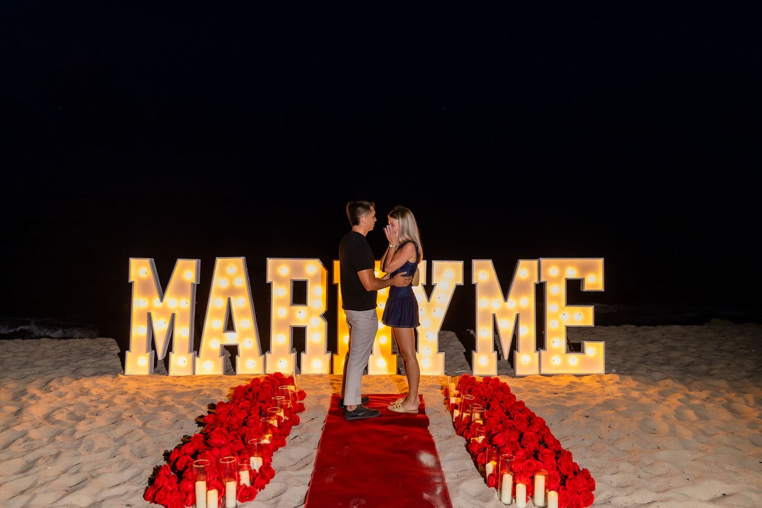 Night beach marriage proposal with glowing MARRY ME letters and red rose petals on sand.
