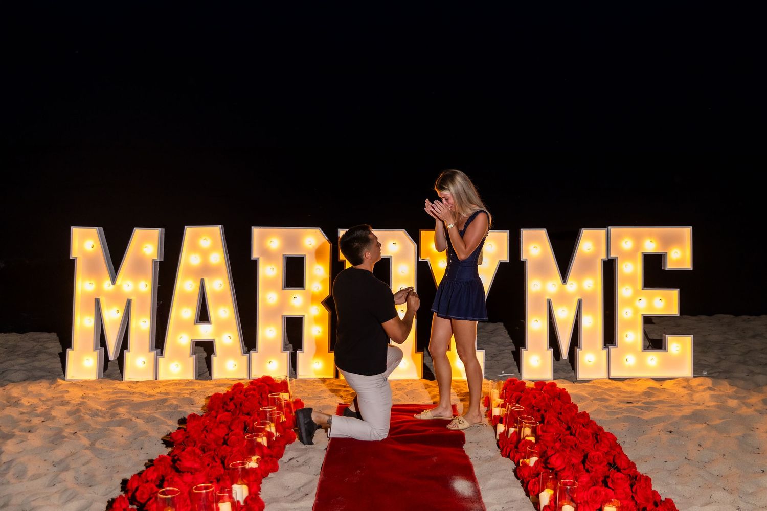 Glowing marquee letters spell out MARRY ME on the beach at night with red roses creating a path.