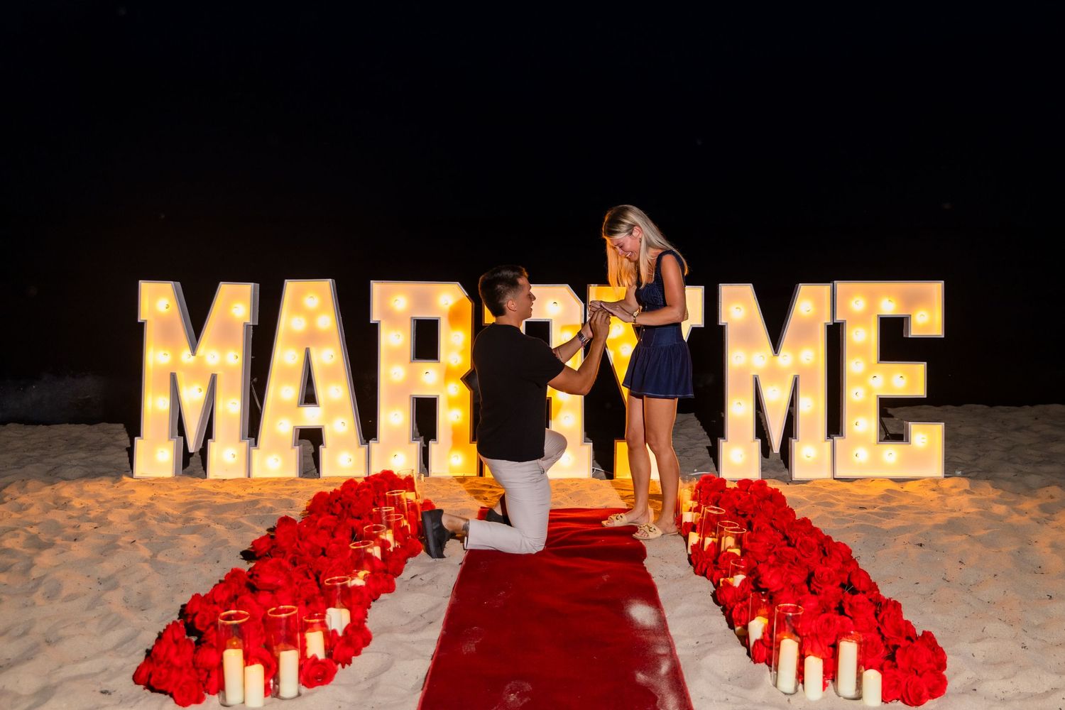 Romantic proposal setup at night with illuminated 'MARRY ME' letters and red rose petals forming a pathway.