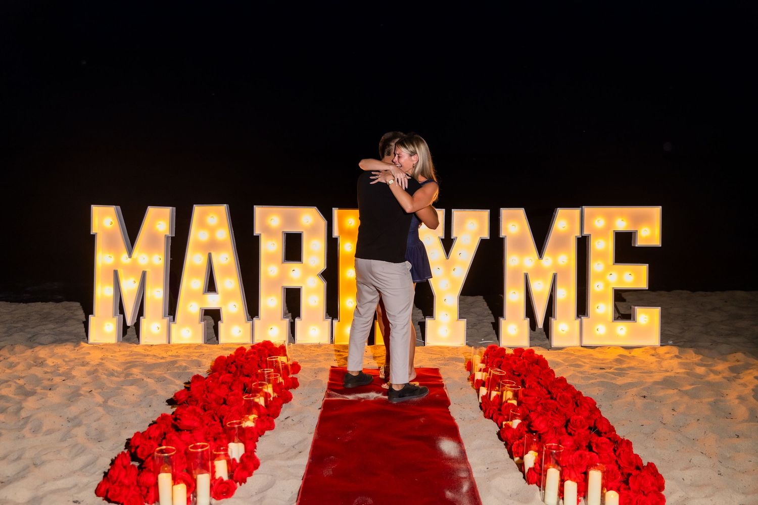 Large illuminated MARRY ME letters set up on a beach at night with red rose petals.
