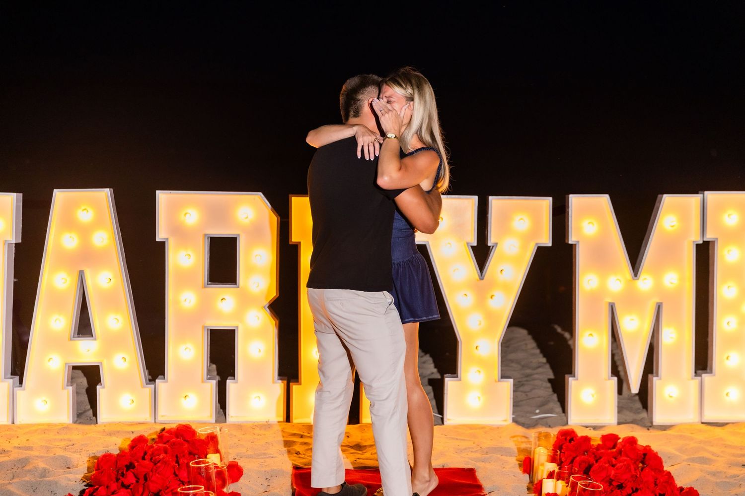 Illuminated marquee letters spell out MARRY ME on a beach with red rose petals lining walkway.