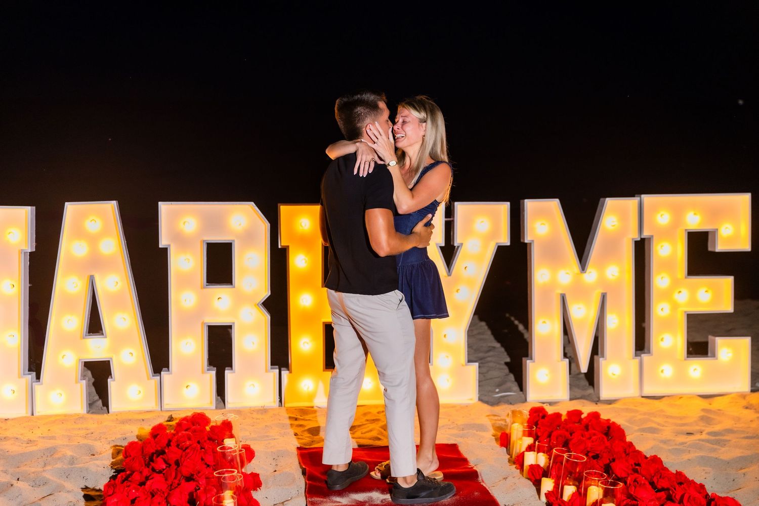 Illuminated marquee letters spell out MARRY ME with red rose petals scattered in front creating a romantic proposal scene.