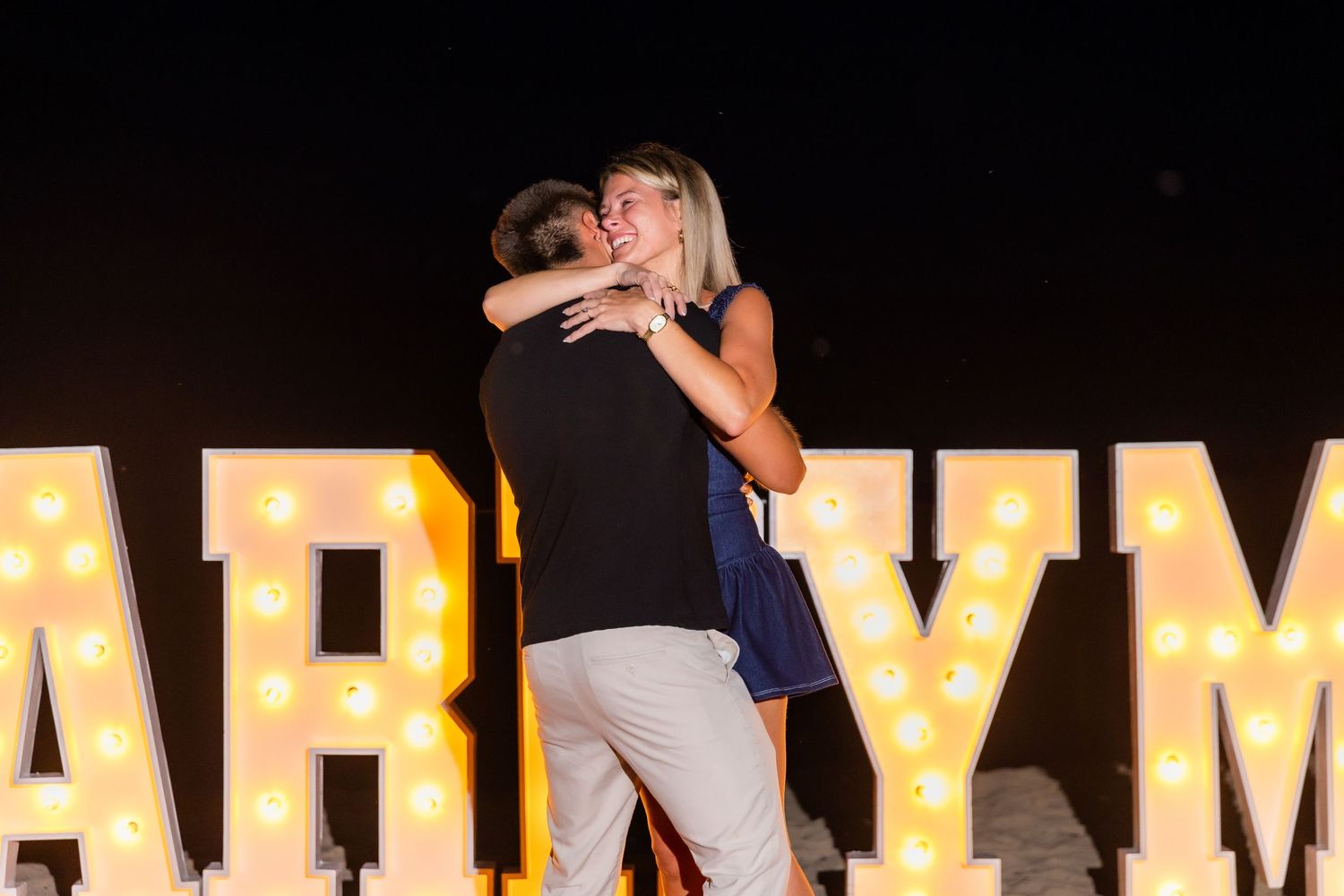 Glowing MARRY ME marquee letters arranged on the beach with decorative lighting.