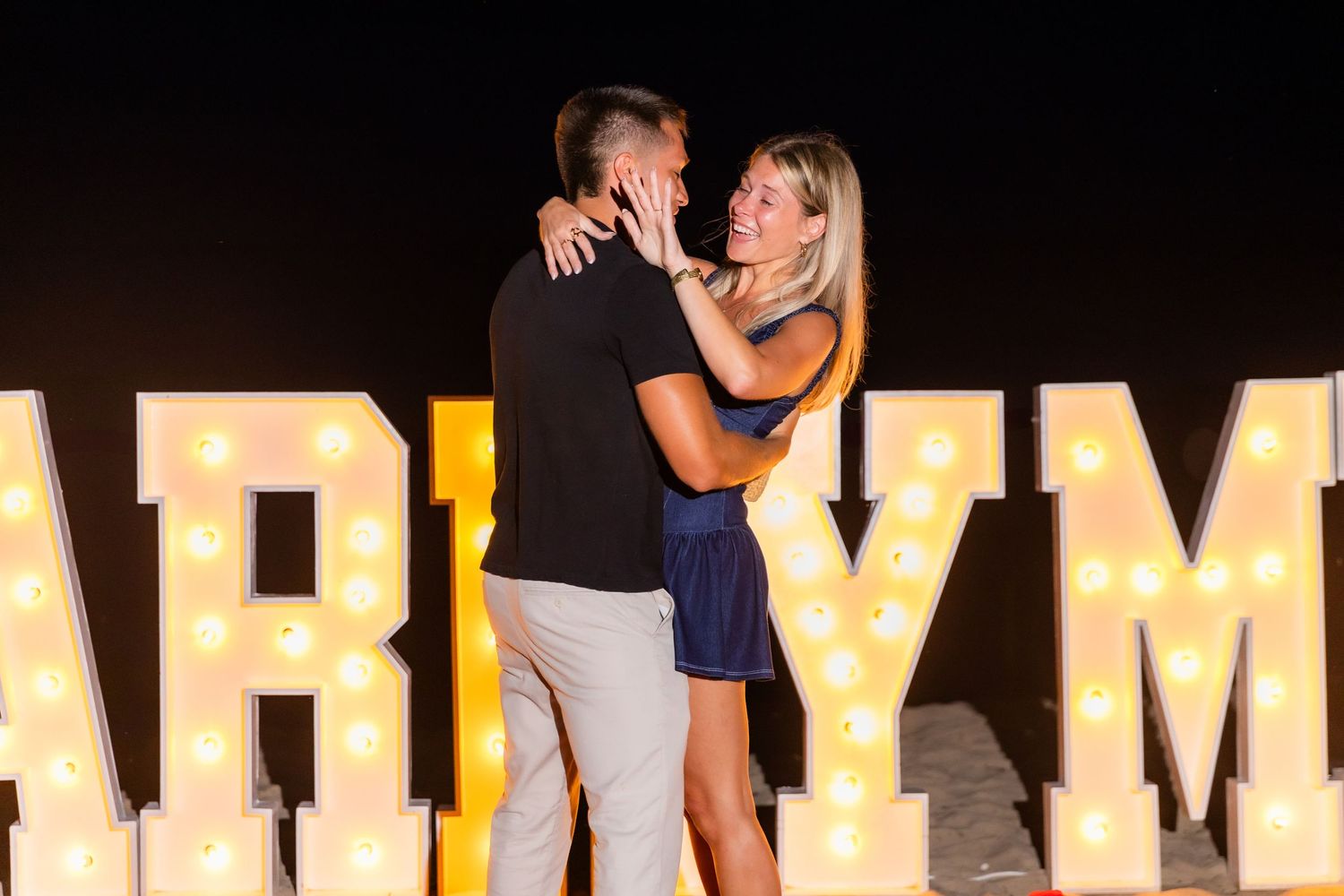 Couple sharing a romantic moment in front of large lit-up MARRY ME letters during a nighttime proposal.