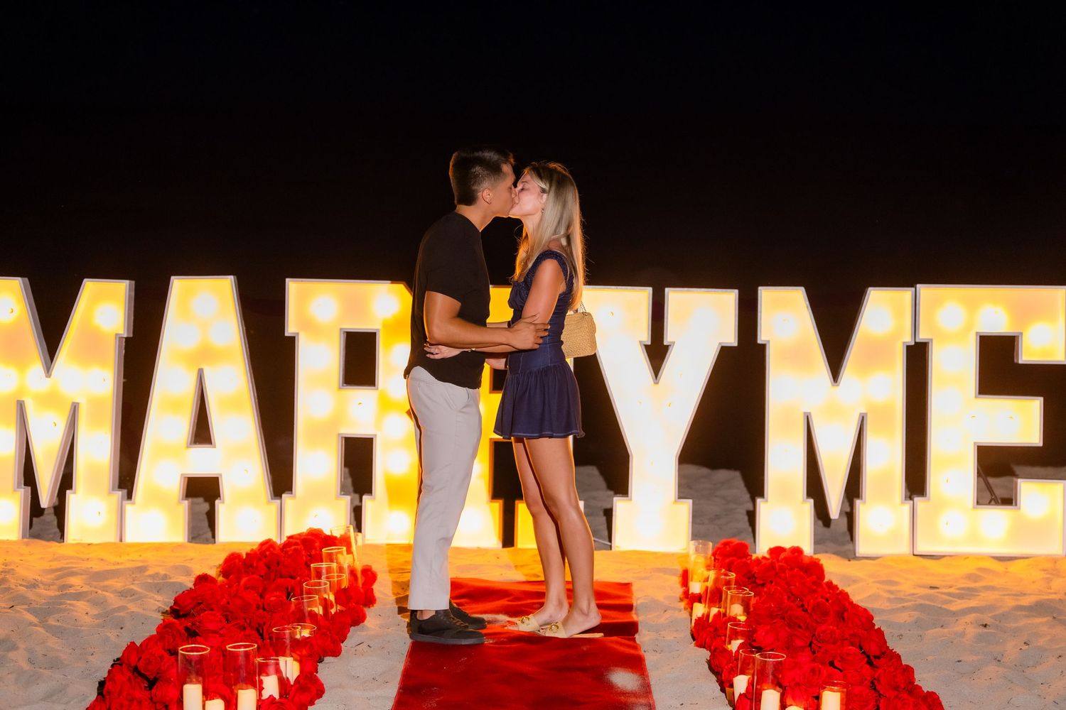 Romantic beach engagement scene with glowing letters and rose petals under night sky.